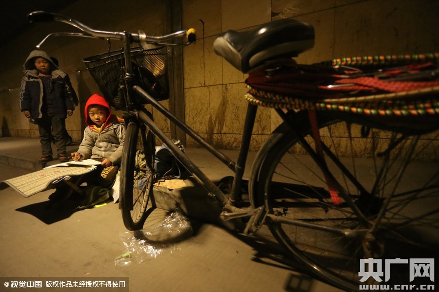 2015年11月8日晚，鄭州，7歲男孩涵洞橋下陪父母練攤，路邊燈下坐小馬扎上寫作業(yè)。